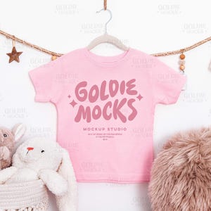 May include: A pale pink t-shirt with the text "GOLDIE MOCKS" in a darker pink, hanging on a grey hanger. The shirt is part of a mockup studio. A plush bunny and a basket are in the lower left corner.