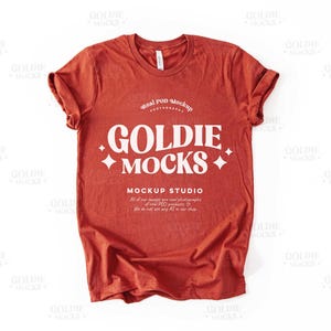 May include: A rust-colored t-shirt with a white graphic that says "Goldie Mocks" and "Mockup Studio". The text is in a retro font style.