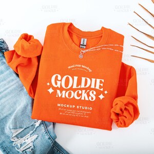 Puede incluir: Sudadera naranja con el texto "GOLDIE MOCKS MOCKUP STUDIO" en blanco. La sudadera está colocada sobre una superficie blanca con un par de pantalones vaqueros azules y una hoja de palma seca.