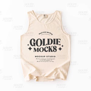 May include: A cream-colored tank top with the text "GOLDIE MOCKS" in large black letters. The tank top has a round neckline and a tag at the back. The words "MOCKUP STUDIO" and other text are also visible.