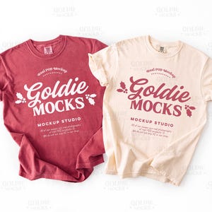 Puede incluir: Se muestran dos camisetas, una burdeos y otra crema. Cada camiseta presenta el texto "Goldie Mocks" en una fuente estilizada, con texto adicional debajo. Las camisetas están dobladas, mostrando el diseño.