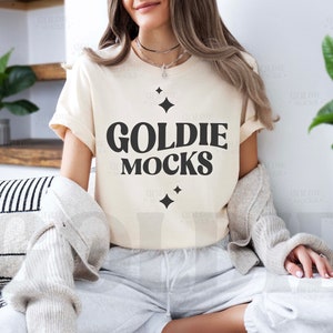 May include: A woman wearing a light beige t-shirt with the text "GOLDIE MOCKS" in black lettering. The shirt has a casual fit and short sleeves. The woman is sitting on a couch with a striped pillow behind her.