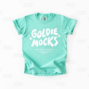 May include: A turquoise t-shirt with rolled-up sleeves. The shirt features the white text "GOLDIE MOCKS" in a playful font, with the words "MOCKUP STUDIO" and additional text below. The shirt is displayed on a white background.