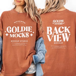 May include: A rust-colored t-shirt with the text "GOLDIE MOCKS" in a white, bold font on the front. The back of the shirt displays "BACK VIEW MOCKUP" in a similar style. The shirt is part of a mockup studio.