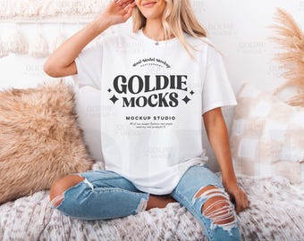 Puede incluir: Una camiseta blanca de gran tamaño con un diseño gráfico negro que dice "Goldie Mocks Mockup Studio". La camiseta la lleva puesta una persona sentada en una manta blanca peluda.