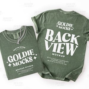 May include: Two olive green t-shirts with white text and a silver chain necklace. One shirt displays "GOLDIE MOCKS" and "MOCKUP STUDIO" text. The other shirt shows "BACK VIEW MOCKUP" text. The shirts are on a white background.