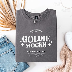Puede incluir: Una camiseta gris con el texto "Goldie Mocks" y "Mockup Studio" impreso en ella. La camiseta está colocada sobre una superficie blanca con un par de zapatos blancos, un par de pantalones vaqueros azules y una manta peluda marrón.