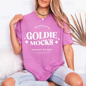 Può includere: Una maglietta rosa con la scritta bianca "GOLDIE MOCKS", con le parole "Real Model Mockup Photography" sopra e "MOCKUP STUDIO" sotto. La maglietta è indossata.