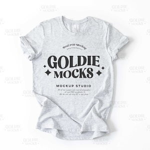 Può includere: Un mockup di una t-shirt grigia con il testo "Goldie Mocks" e "Mockup Studio" stampato sul davanti. Il testo è in un font retrò nero.