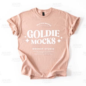 May include: A light peach colored t-shirt with a white graphic that says "Goldie Mocks" and "Mockup Studio".