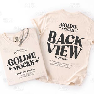 May include: Two cream-colored t-shirts, one folded and one displayed from the back. The folded shirt has a silver chain necklace and the text "GOLDIE MOCKS" and "MOCKUP STUDIO". The back of the other shirt reads "BACK VIEW MOCKUP".