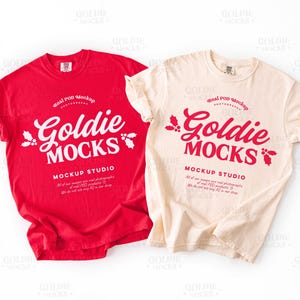 May include: Two t-shirts, one red and one cream-colored, are displayed. Each shirt features the text "Goldie Mocks" in a stylized font, along with additional text. The shirts are folded at the sleeves and are on a white background.