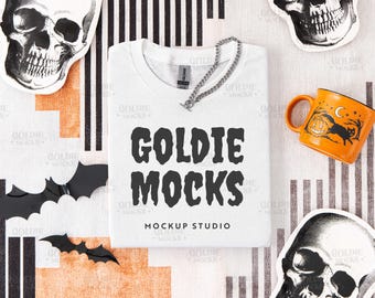 May include: A white t-shirt with the text "GOLDIE MOCKS" in black, a silver chain, and an orange mug with a Halloween design. The image also includes skull stickers and black bat cutouts, creating a spooky theme.