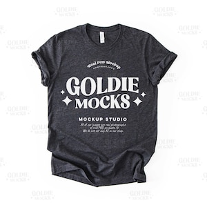 Könnte beinhalten: Ein dunkelgraues T-Shirt mit einer weißen Grafik, auf der "Goldie Mocks" und "Mockup Studio" steht. Die Grafik enthält auch eine kleine Diamantenform auf jeder Seite des Textes.