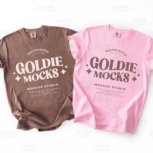 Może przedstawiać: Dwie koszulki, jedna brązowa i jedna różowa, są prezentowane na białym tle. Każda koszulka ma napis "GOLDIE MOCKS" w stylizowanej czcionce, z dodatkowym tekstem poniżej. Koszulki są casualowe i wydają się być wykonane z miękkiego materiału.