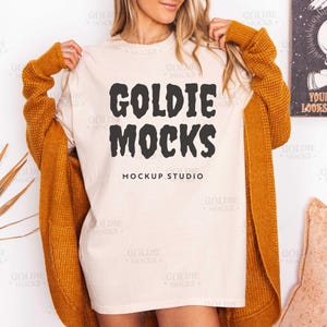 May include: A cream-colored t-shirt with the text "GOLDIE MOCKS" in a bold, black font. The words "MOCKUP STUDIO" are printed below. The shirt is worn with a mustard-colored cardigan. A decorative dried plant and a pillow are in the background.