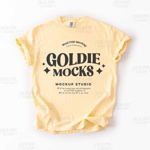 以下が含まれることがあります： レトロなフォントで「Goldie Mocks」というテキストが書かれた黄色のTシャツのモックアップです。「Real POD Mockup Photography」というテキストも、より小さなフォントで表示されています。「Mockup Studio」というテキストはロゴの下にあります。「All of our images are real photographs of real POD products. We do not use any AI in our shop.」というテキストはスタジオ名の下にあります。