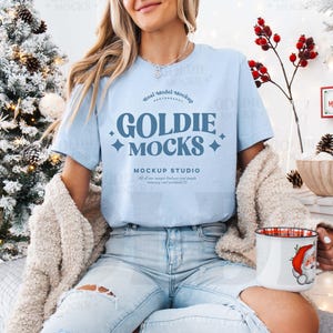 May include: A light blue t-shirt with the text "Goldie Mocks Mockup Studio" in a blue and white font. The shirt is being worn by a person with long blonde hair.