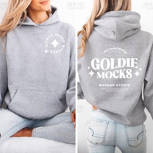 Puede incluir: Una sudadera con capucha gris jaspeado con un bolsillo delantero y una capucha. La parte delantera de la sudadera tiene un gráfico circular blanco con texto. La parte trasera de la sudadera tiene el texto "GOLDIE MOCKS" en blanco. La sudadera es parte de un estudio de maquetas.