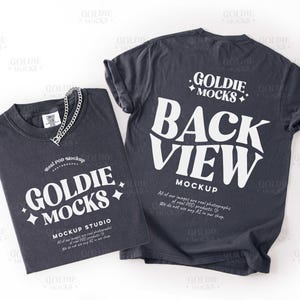 May include: Two dark gray t-shirts with white text. One shirt is folded, displaying "GOLDIE MOCKS MOCKUP STUDIO" and a silver chain. The other shirt shows "BACK VIEW MOCKUP" in large letters. Both shirts have additional text.