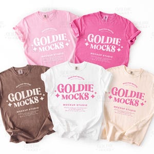 May include: Five different colored t-shirts with the text "Goldie Mocks" and "Mockup Studio" printed on them. The shirts are laid out on a white surface.