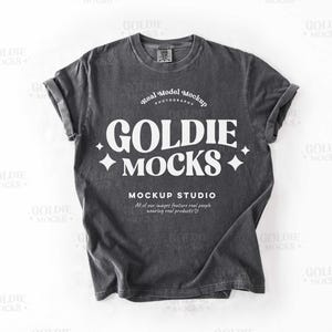 Puede incluir: Una camiseta gris oscura con un gráfico blanco que dice "GOLDIE MOCKS" y "MOCKUP STUDIO". El gráfico está en una fuente de estilo retro.