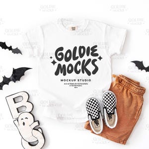 May include: A white t-shirt with the text "GOLDIE MOCKS" and a pair of black and white checkered slip-on shoes, orange shorts, and Halloween decorations, including bats and a ghost.