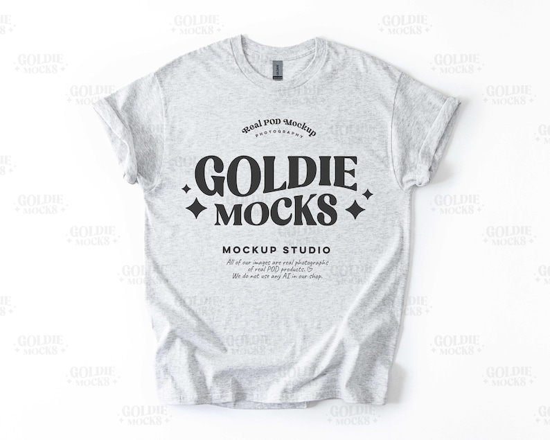 May include: A gray t-shirt mockup with the text "Goldie Mocks" in a black retro font. The text is surrounded by small diamond shapes. The text "Real Pod Mockup Photography" is above the main text and the text "Mockup Studio" is below the main text. The text below the main text reads "All of our images are real photographs of real POD products. We do not use any AI in our shop."