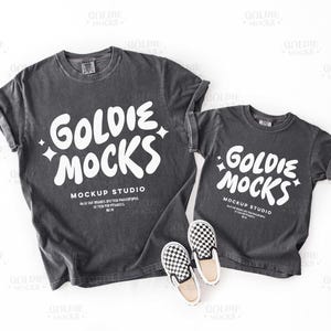 May include: Two charcoal gray t-shirts, one adult and one child size, with the text "Goldie Mocks" in white. The adult shirt is next to a pair of black and white checkered slip-on shoes. Mockup Studio text is also visible.