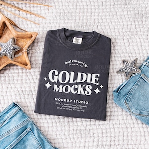 Może przedstawiać: Czarny T-shirt z napisem "GOLDIE MOCKS" w białych literach. Koszulka leży płasko na białym, puszystym materiale z niebieskimi dżinsami i ozdobą w kształcie gwiazdy.
