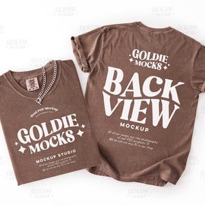 以下が含まれることがあります： 白文字とシルバーチェーンネックレスが付いた2枚の茶色のTシャツ。1枚のシャツには「GOLDIE MOCKS」と「MOCKUP STUDIO」と表示されています。もう1枚のシャツには「BACK VIEW MOCKUP」と表示されています。シャツは折りたたんで平らに置かれています。