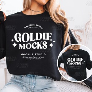 Puede incluir: Una sudadera negra con un diseño gráfico blanco que dice "Goldie Mocks" y "Mockup Studio". El diseño gráfico está en una fuente de estilo retro. La sudadera está siendo usada por una modelo.