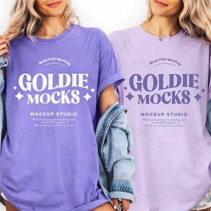 Puede incluir: Dos mujeres con camisetas moradas y lavanda de gran tamaño. Las camisetas tienen el texto "GOLDIE MOCKS" en una fuente grande y estilizada, con "MOCKUP STUDIO" debajo. Las camisetas son de color liso con un aspecto vintage.