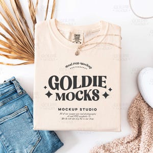 Puede incluir: Una camiseta beige de cuello redondo con el texto "Goldie Mocks" y "Real Pod Mockup" impreso en la parte delantera. El texto está en negro, en negrita. La camiseta está colocada sobre una superficie blanca con un par de zapatillas blancas, unos vaqueros azules y una manta de punto beige.