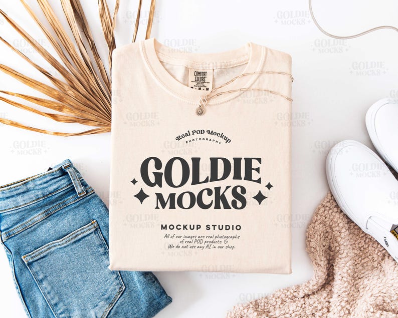 May include: A beige crew neck t-shirt with the text "Goldie Mocks" and "Real Pod Mockup" printed on the front. The text is in a black, bold font. The t-shirt is laying flat on a white surface with a pair of white sneakers, a pair of blue jeans, and a beige knitted throw.