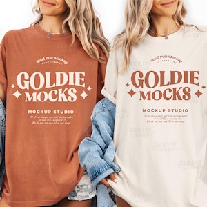Puede incluir: Se muestran dos camisetas, una de color óxido y otra crema. Cada camiseta presenta el texto "GOLDIE MOCKS" en una fuente retro, con texto adicional debajo. Las camisetas forman parte de un estudio de maquetas.