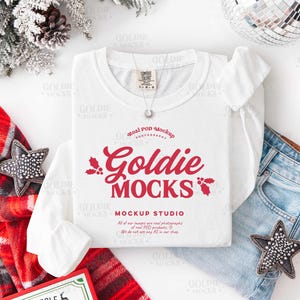 May include: A white long-sleeve t-shirt with the text "Goldie Mocks" in red script, a mockup studio logo, and a small silver necklace. The shirt is paired with blue jeans and festive holiday decorations.
