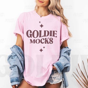 May include: A pink t-shirt with the text "Goldie Mocks" in brown lettering and two small stars on either side of the text. The shirt is being worn by a person with long blonde hair and a denim jacket.