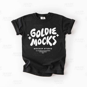 May include: Black t-shirt with rolled sleeves. The shirt features the white text "GOLDIE MOCKS" in a playful font, with the words "MOCKUP STUDIO" below. The shirt is a mockup for apparel.