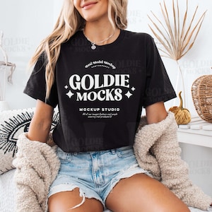 以下が含まれることがあります： 白いソファに座っている女性。黒いTシャツを着ており、白い文字で「Goldie Mocks Mockup Studio」と書かれています。デニムのショートパンツを履いています。白い毛布がかけられています。
