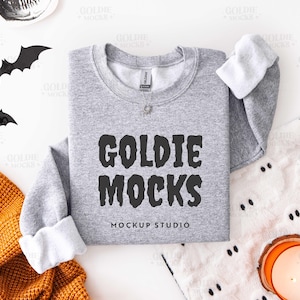 Op de afbeelding: Een grijze sweatshirt met de tekst "GOLDIE MOCKS MOCKUP STUDIO" in het zwart. De mouwen zijn opgerold en tonen witte stof. Decoratieve vleermuizen en een kaars staan op de achtergrond.