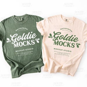 May include: Two short-sleeved t-shirts, one olive green and one cream, are displayed. Both shirts feature the text "Goldie Mocks" in a stylized font, with holly leaf accents. The shirts are laid flat on a white surface.