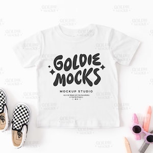 Könnte beinhalten: Weißes T-Shirt-Mockup mit dem Text "Goldie Mocks Mockup Studio" in Schwarz. Das Shirt liegt flach auf einer weißen Oberfläche mit einem Paar schwarz-weiß karierter Slipper und bunten Buntstiften.