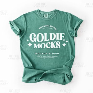 May include: A teal green t-shirt with a white graphic that says "GOLDIE MOCKS" and "MOCKUP STUDIO" with a small star design. The shirt is folded and laid flat on a white surface.