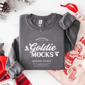 May include: A gray sweatshirt with the text "Goldie Mocks" in white, a silver necklace, and a Christmas-themed gift bag. The sweatshirt is part of a mockup studio setup, with holiday decorations.