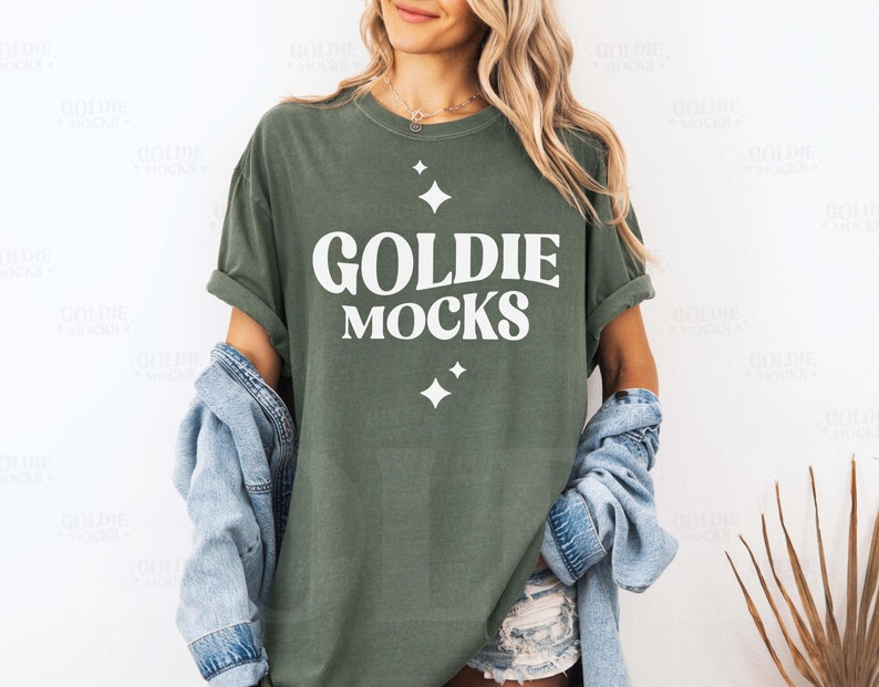 Op de afbeelding: Olijfgroen t-shirt met de witte tekst "GOLDIE MOCKS" en twee witte sterrenburst-afbeeldingen. Een lichtblauwe spijkerjas is over de schouders gedrapeerd. Het t-shirt heeft opgerolde mouwen.