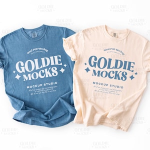 Könnte beinhalten: Zwei T-Shirts, eines in Blau und eines in Beige, werden gezeigt. Jedes Shirt trägt den Text "GOLDIE MOCKS" in einer stilisierten Schrift, darunter "MOCKUP STUDIO". Die Shirts liegen flach, um ihr Design zu präsentieren.