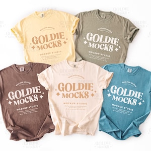 Könnte beinhalten: Fünf T-Shirts in verschiedenen Farben mit dem Aufdruck "Goldie Mocks" und "Mockup Studio". Die Shirts liegen auf einer weißen Oberfläche.
