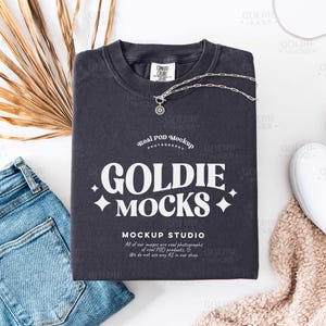 Puede incluir: Una composición plana con una camiseta gris oscuro que muestra el texto "GOLDIE MOCKS" y un collar plateado. La camiseta se combina con vaqueros azul claro, zapatos blancos sin cordones y una hoja de palma seca. La imagen incluye el texto "Real POD Mockup PHOTOGRAPHY".