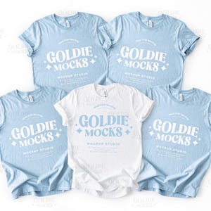 Puede incluir: Cinco camisetas en una disposición plana. Cuatro son de color azul claro y una es blanca. Todas tienen el texto "Goldie Mocks" en una fuente retro con estrellas a cada lado. El texto "Mockup Studio" está debajo del texto principal.
